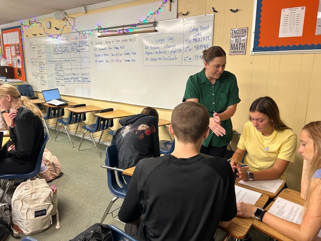 A classroom with desks; a teacher wearing a green shirt stands between two students. Four other students are seated.