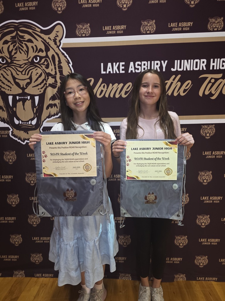 Two girls hold certificates in front of a tiger image and school name on a dark backdrop.
