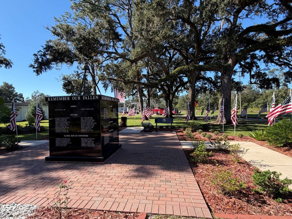 A memorial with names listed on a wall, surrounded by trees and flags. A paved pathway leads to the memorial.