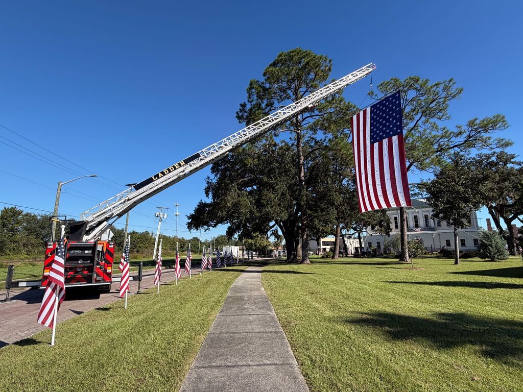 Fire truck with extended ladder holds flag. American flags on grass. Trees and building in background.