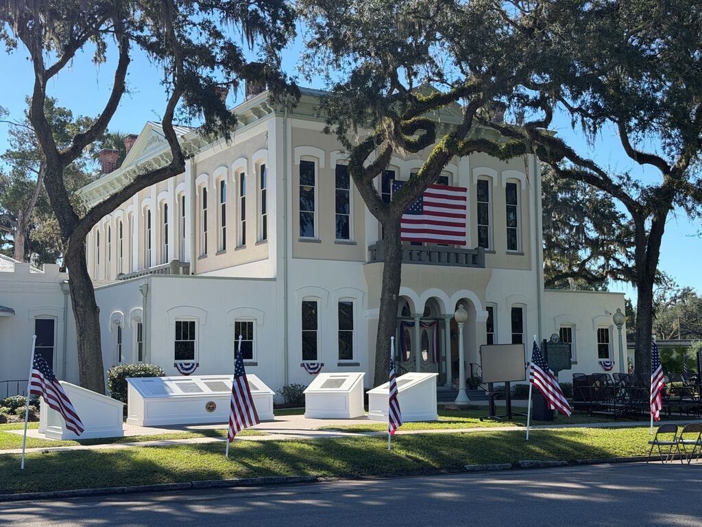 A white building with multiple windows, an American flag on the balcony, and American flags on display on the front lawn.