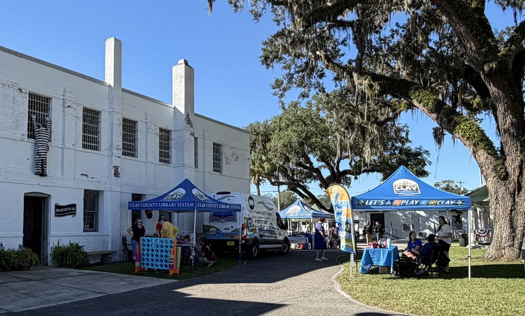 A white building with multiple windows, blue tents, people, and a large tree in the foreground.