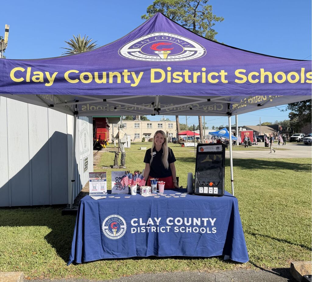 A tent with Clay County District Schools written on it. A woman stands behind a table with a blue cloth.