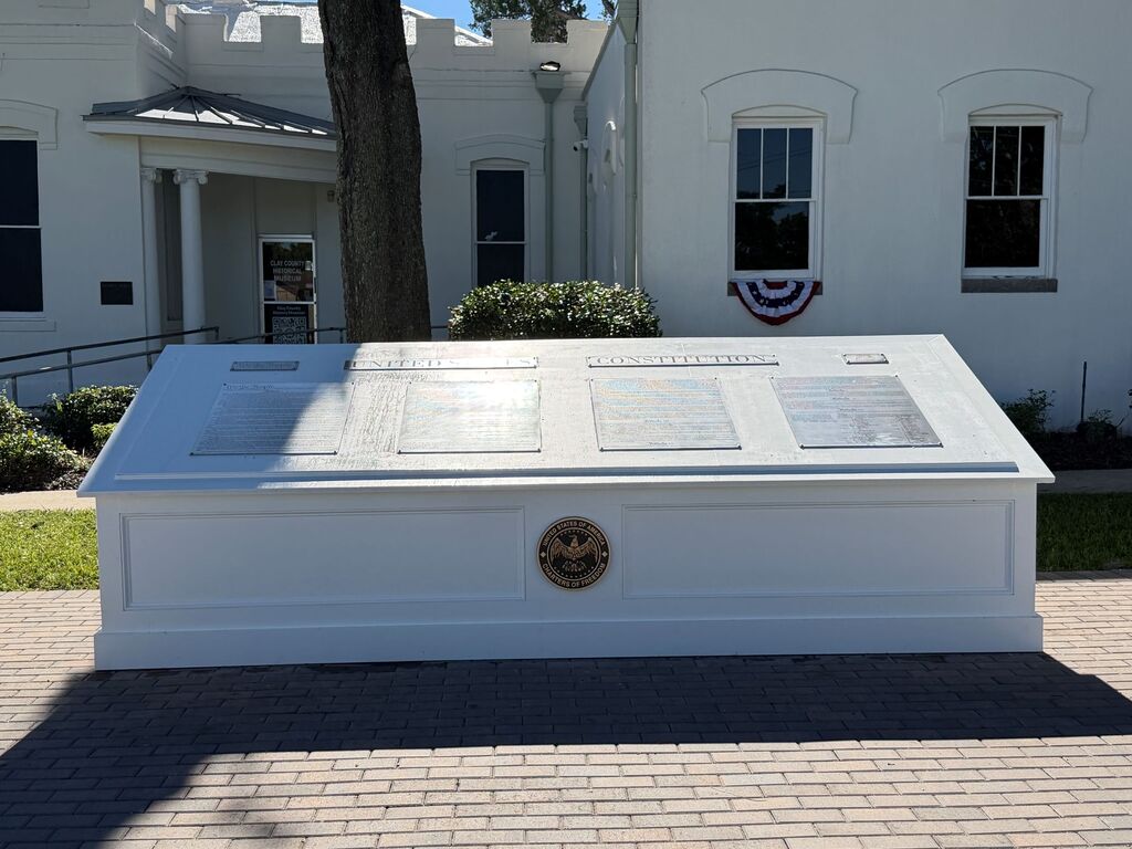 A white box-like monument with several engraved panels and a round emblem in the center, situated in front of a white building.