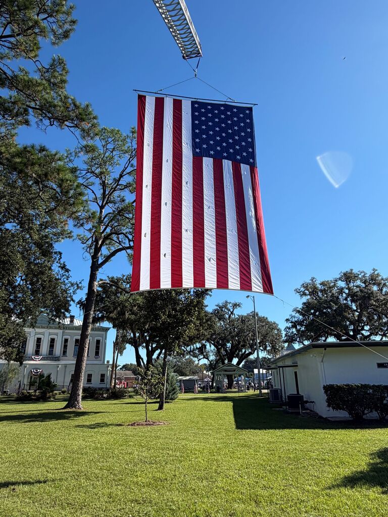 A large American flag suspended by a crane over a grassy area with trees and buildings in the background.