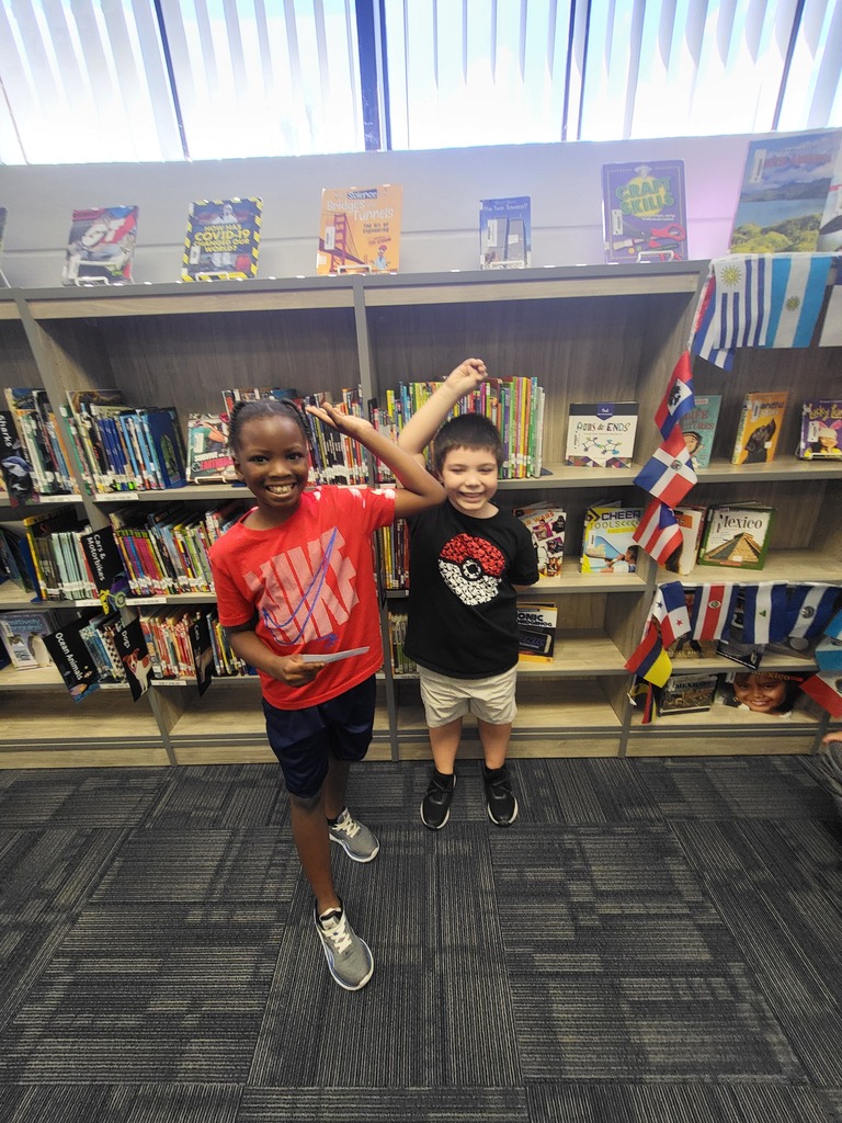 Two kids stand in a library with bookshelves and flags in the background. One holds a paper.