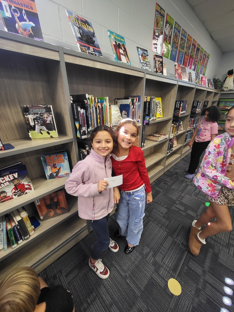 Two girls stand in a library with books on shelves. One holds a piece of paper.