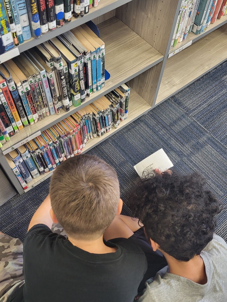 Two children sit on the floor, one reading a book, next to a bookshelf filled with books.