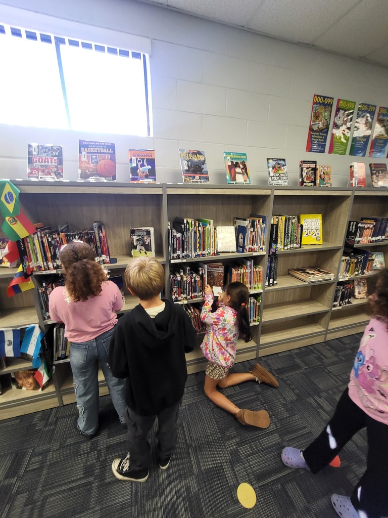 Children browse library books, standing and sitting on the floor in a room with white walls.
