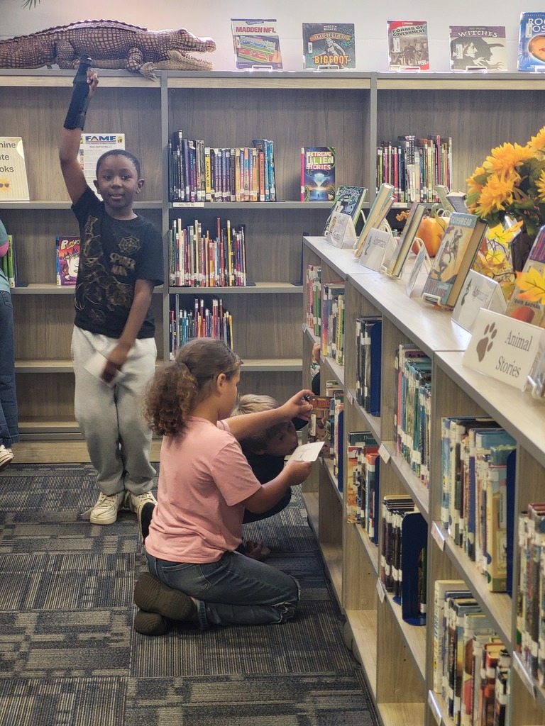 Children in a library, one raising a book and another sitting with a book. Shelves filled with books and plants.