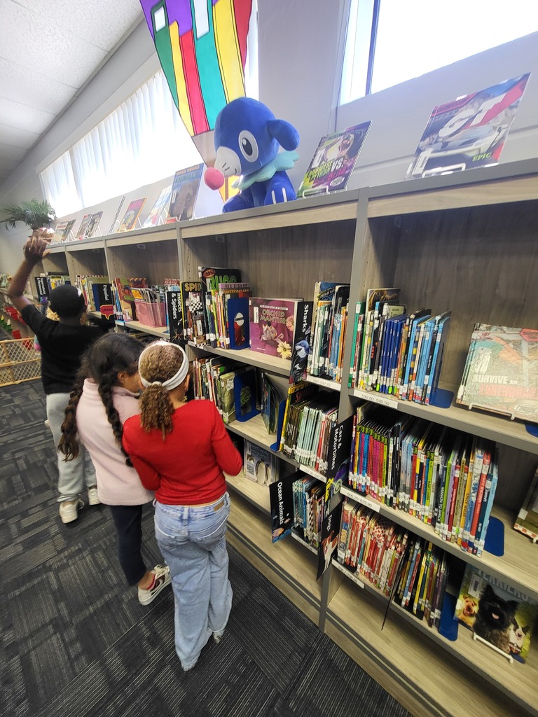 Three children browse a library with tall shelves, books, and a kite decoration. One child reaches for a book.