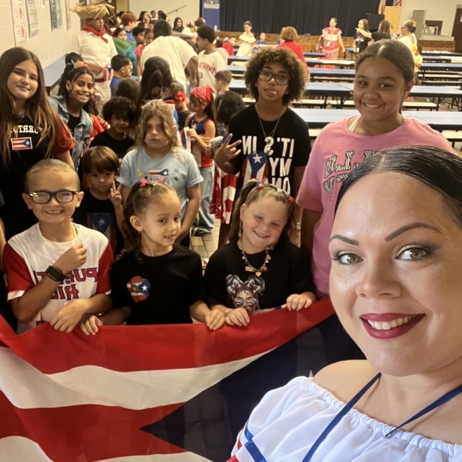 A group of children and adults holding a Puerto Rican flag, standing in a room with tables in the background.