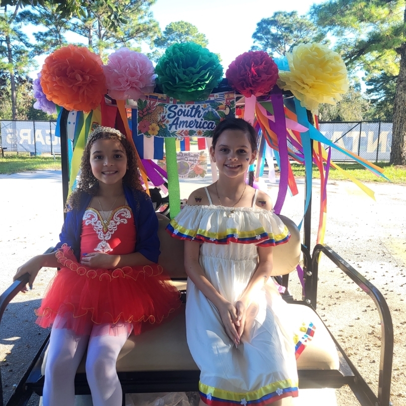 Two girls in costumes on a golf cart. One wears a red tutu, the other a white dress.