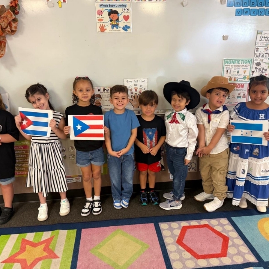 Several children in a classroom hold flags while wearing themed outfits. The carpet has various geometric shapes.