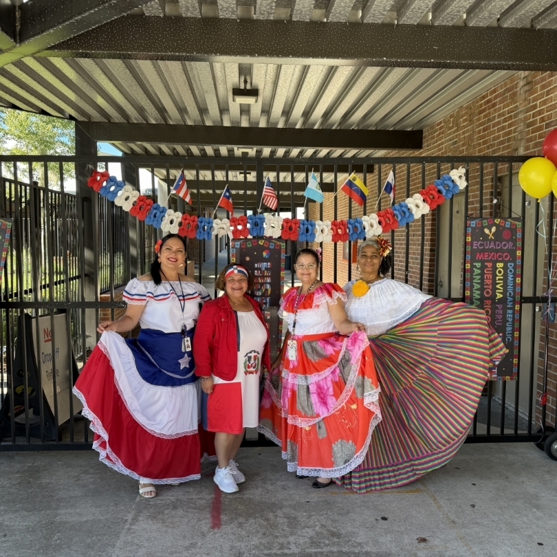 Four women dressed in colorful traditional clothing stand together in front of a gate. Flags hang behind them.
