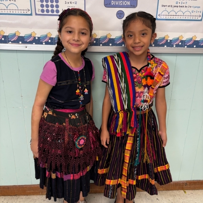 Two girls in colorful dresses stand in a classroom, smiling and standing next to each other.