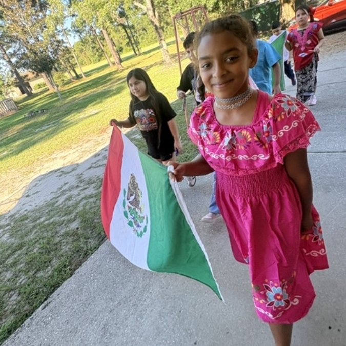 A girl in a pink dress waves a Mexican flag while walking with other children in a park.
