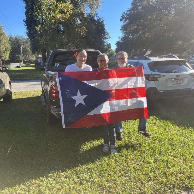 Three people stand on grass, holding a Puerto Rico flag, with a pickup truck and parked cars behind them.