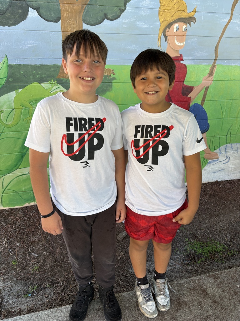 Two boys, both wearing white t-shirts, stand in front of a mural featuring a character and a tree.
