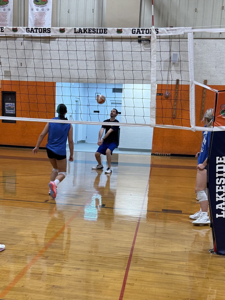 Players practice volleyball with a net in a gym. Two people are on each side. One player runs towards the net.