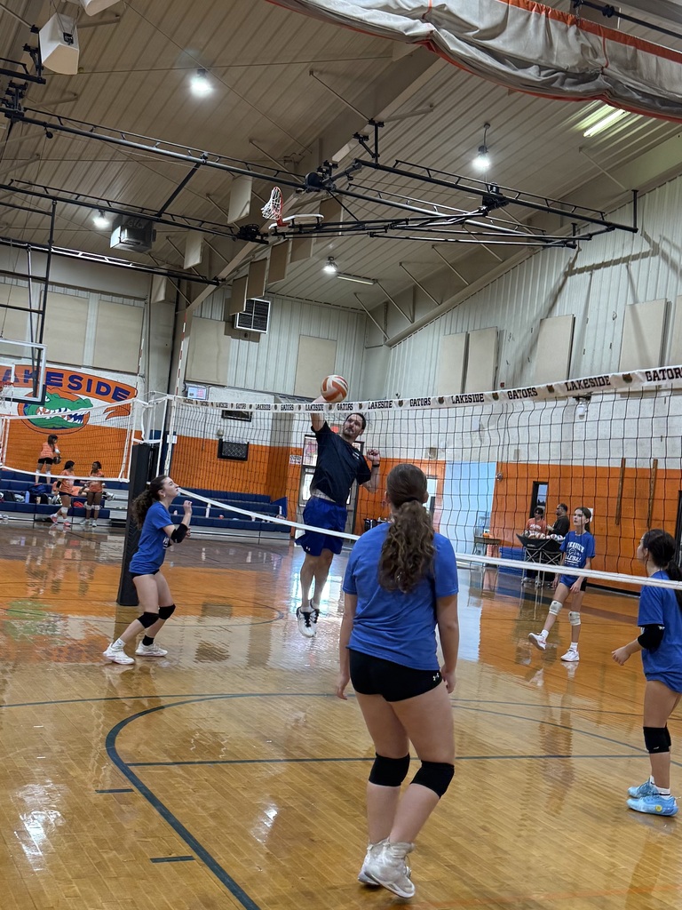 An indoor volleyball game in progress. Players in blue uniforms move across the court. A player leaps to spike the ball.