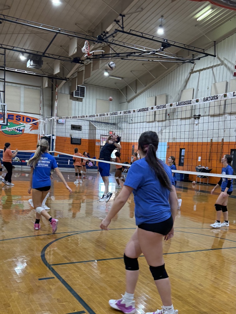 An indoor court with a net and people playing volleyball. Some wear blue shirts and black shorts.