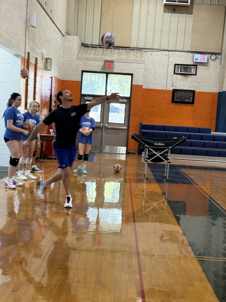 A man in a black shirt and blue shorts throws a volleyball in a gym with kids.