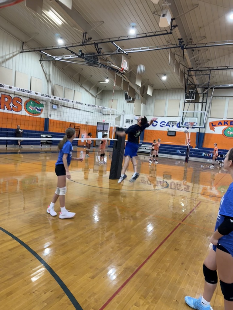 A group of people playing volleyball in a gym. One person jumps to spike the ball.