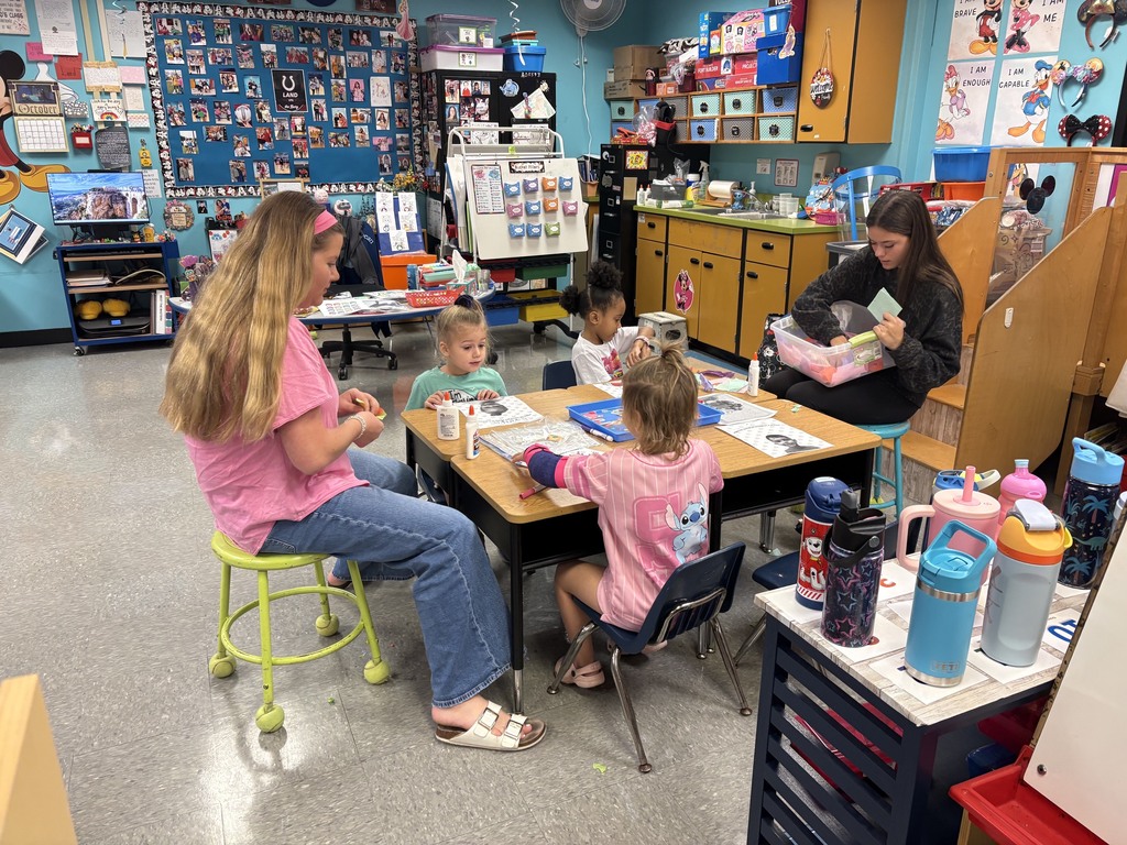 A classroom with colorful walls. Adults and children work at tables, one reading to children.