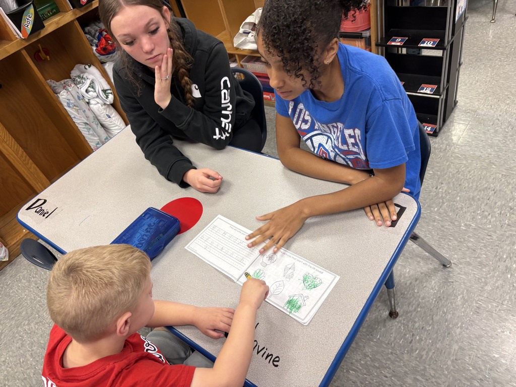 A teacher and student work on a table, with a boy at the table and a girl leaning over.