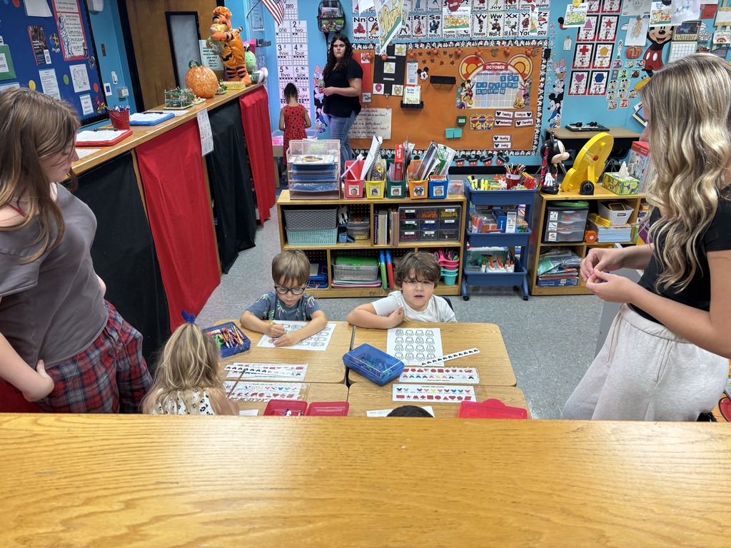 Several children and adults in a classroom setting. Children sit at tables with books and papers.