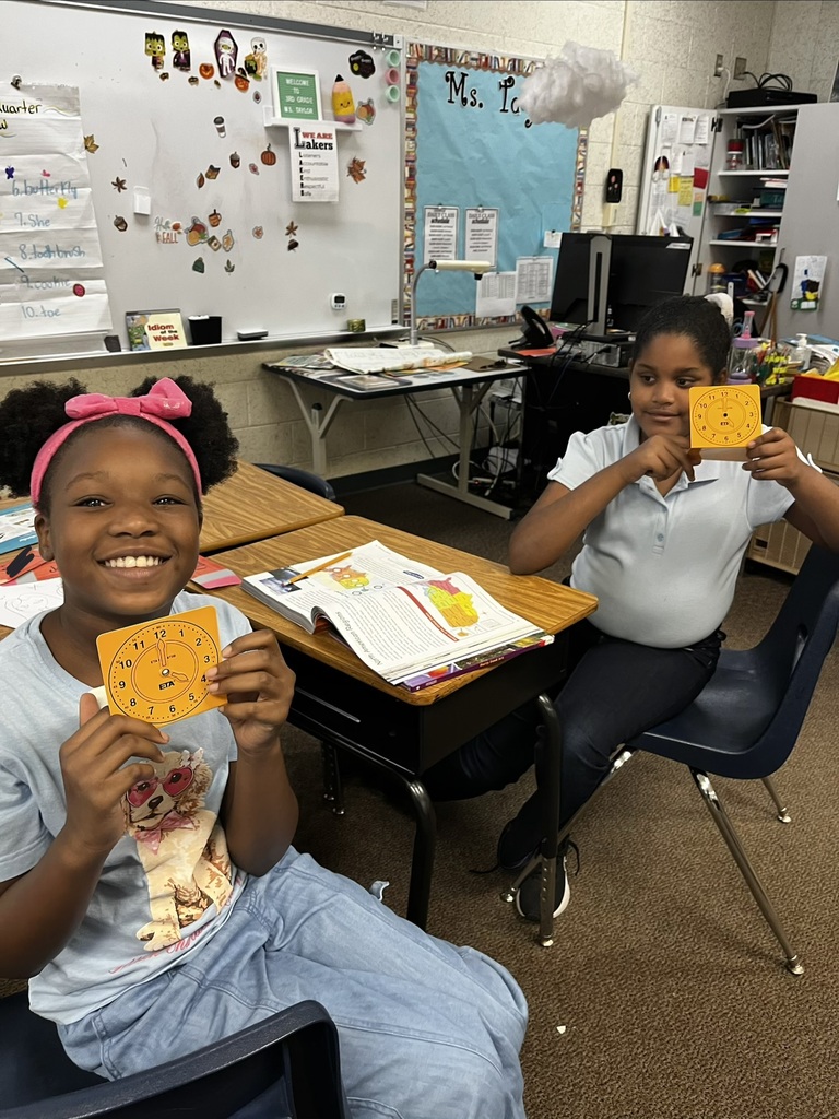Two young girls hold up pieces of paper with clocks. One sits in a chair, the other at a desk.