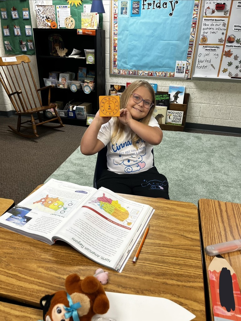 A child in a classroom sits at a desk, holding a small yellow square, with a book and stuffed toy nearby.