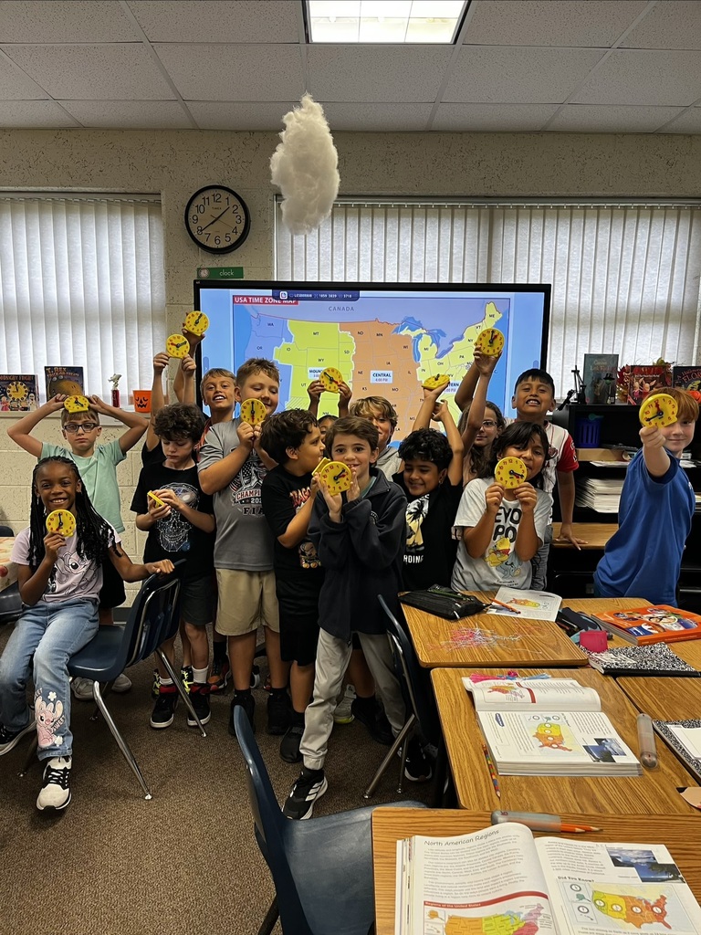 Group of children posing with yellow cutouts in a classroom. Tables with books and a large screen behind.