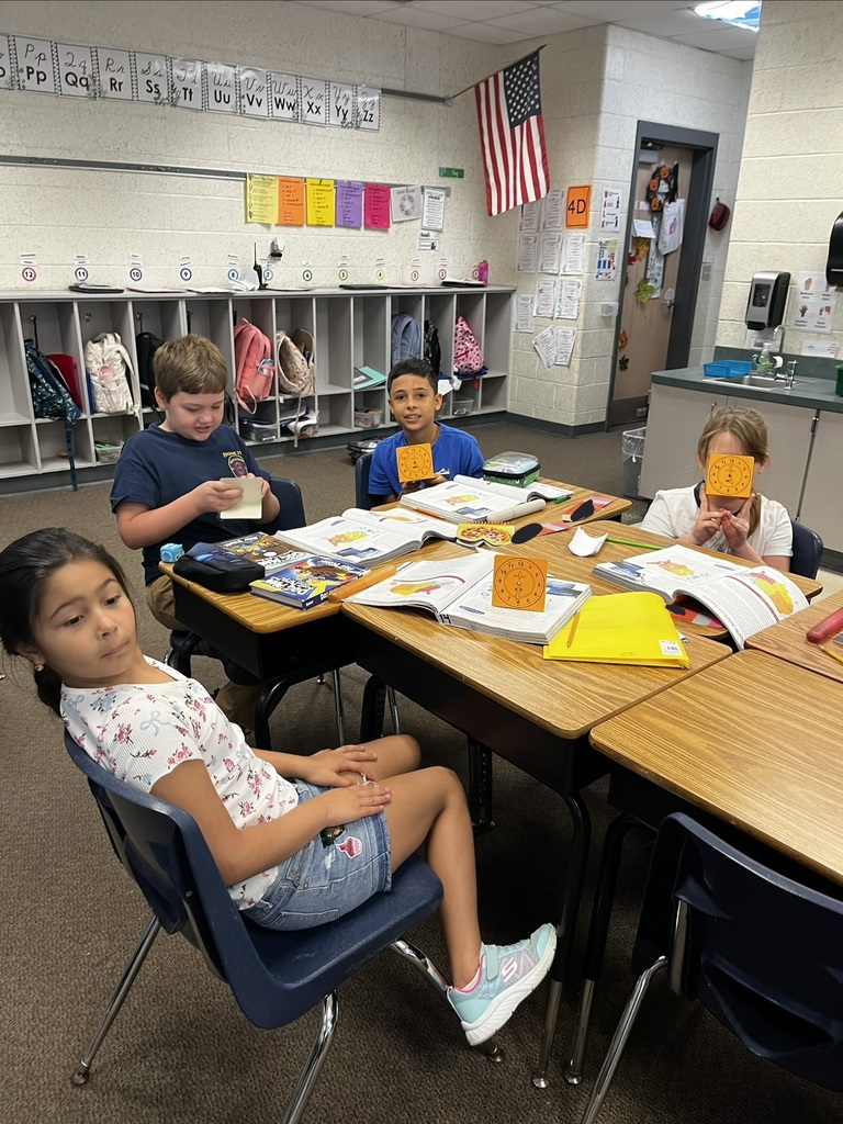 Four children in a classroom setting, sitting at desks. One girl looks down, another reads, and two boys are engaged in discussion.