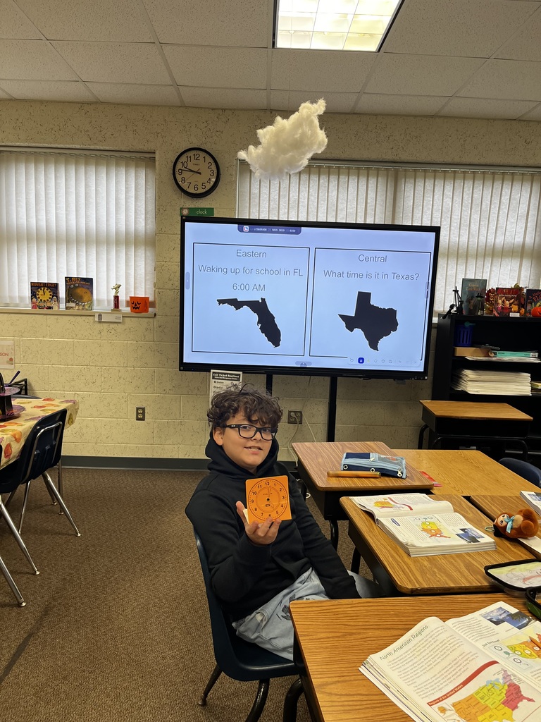 A person sits at a desk in a classroom. The person holds a small card. A large screen displays maps of Florida and Texas.