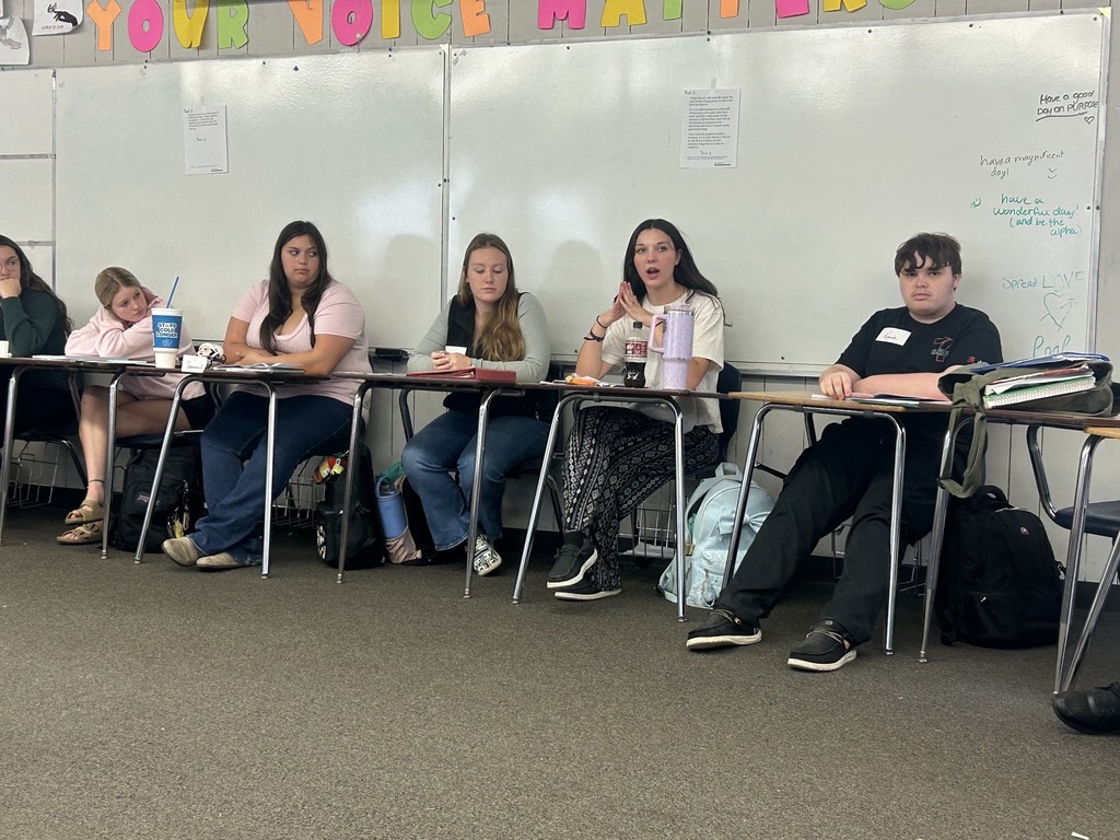 A group of people sit at tables in a classroom. A whiteboard is behind them with colorful letters.