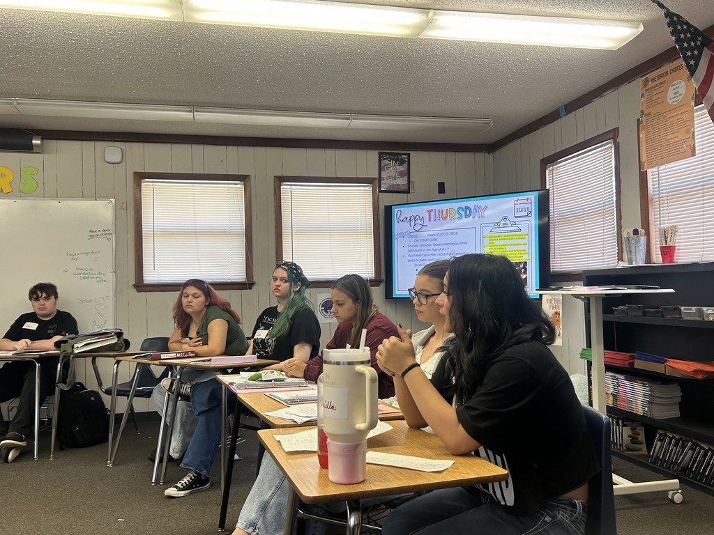 A group of people are seated around tables in a classroom. A large screen is on the wall.