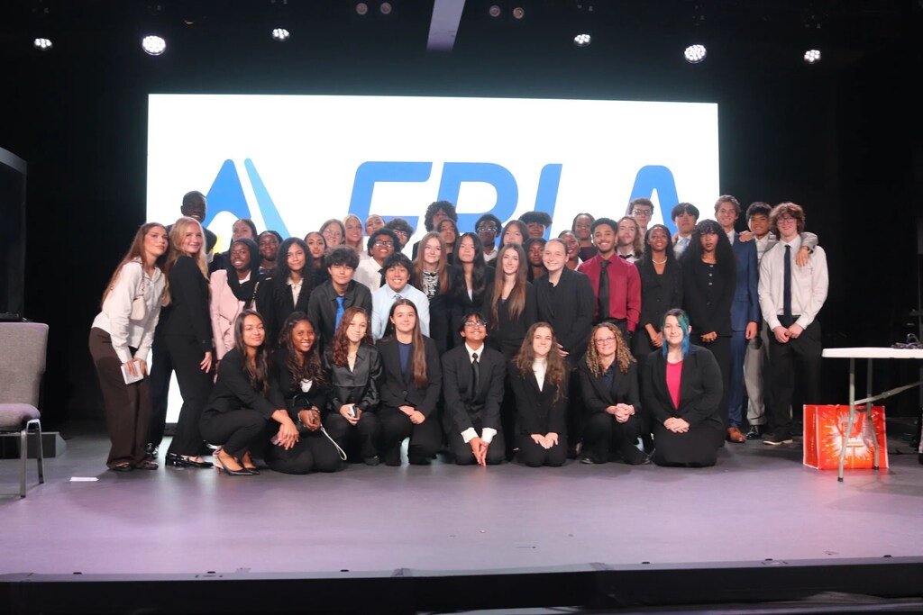 A group of people on a stage, some sitting and some standing, with a large screen in the background.