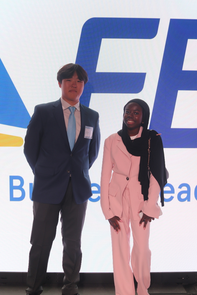Two individuals stand before a screen with a blue logo. The man wears a dark suit and tie; the woman, a pink suit and scarf.