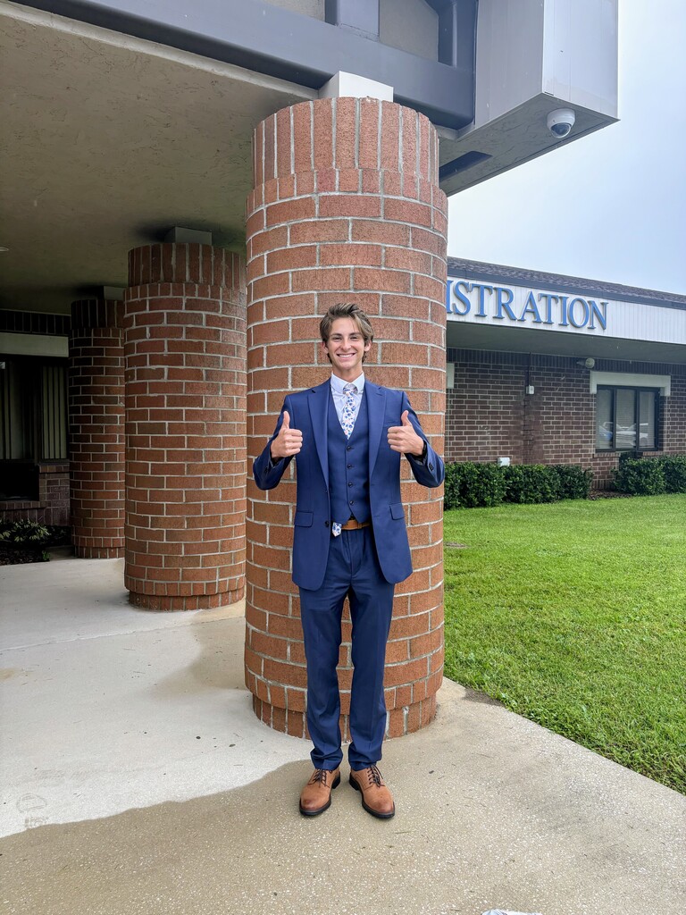 A person stands in a suit with thumbs up, next to a brick column at a school.