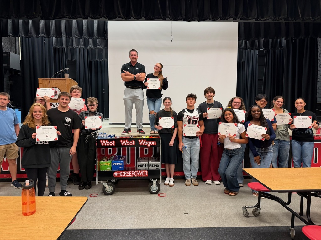 Group of students and adults holding certificates. Some standing on a platform, others in a room with tables.