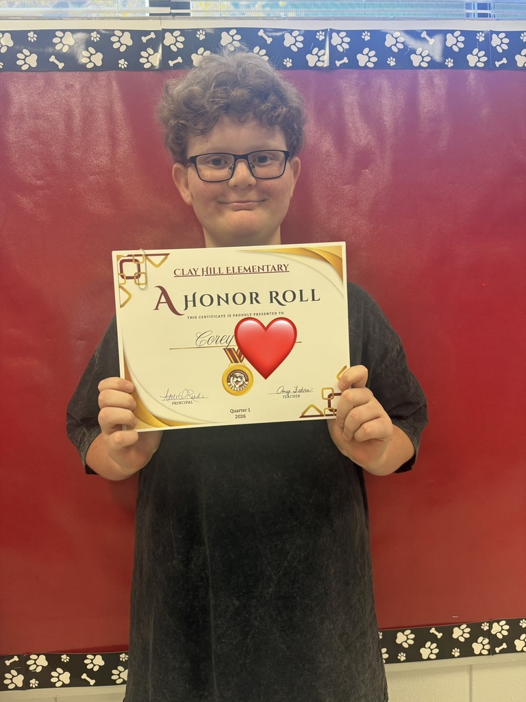 A child wearing glasses and a black shirt holds a certificate with a red heart, standing in front of a red backdrop.