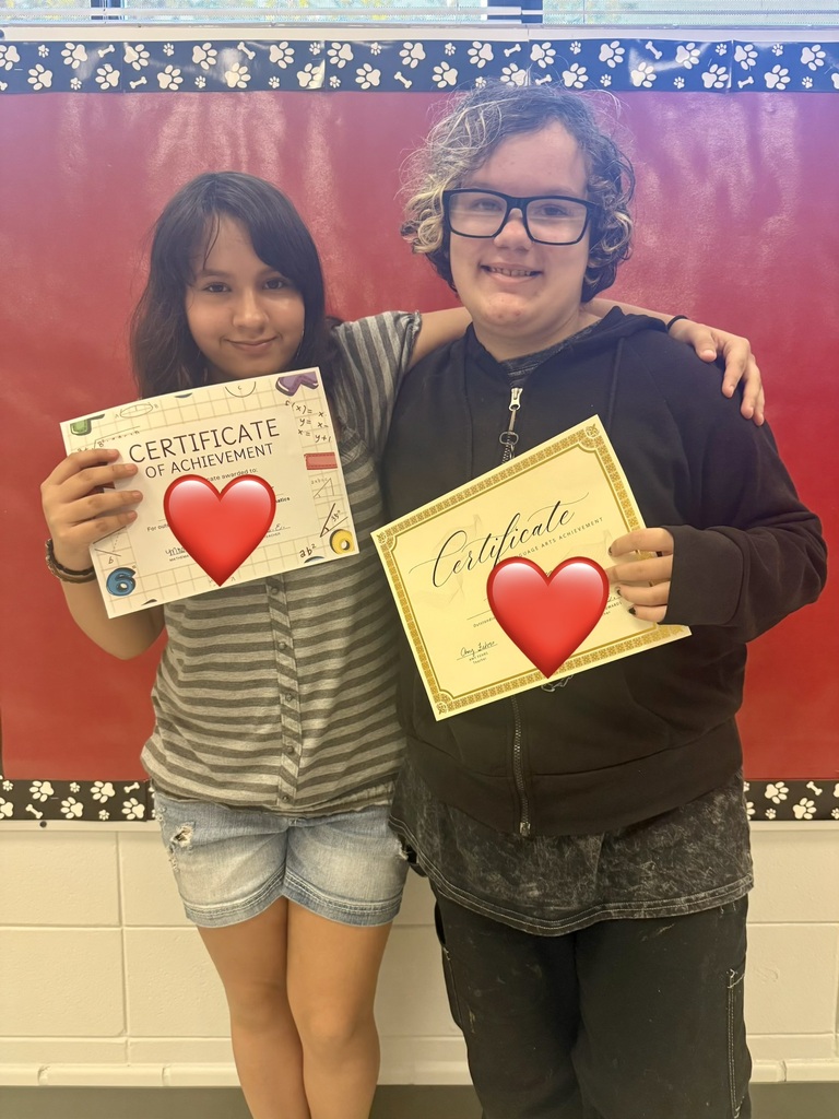 Two individuals stand together, holding certificates with heart graphics. One has a striped shirt and denim shorts, the other wears a black jacket. Background is red.