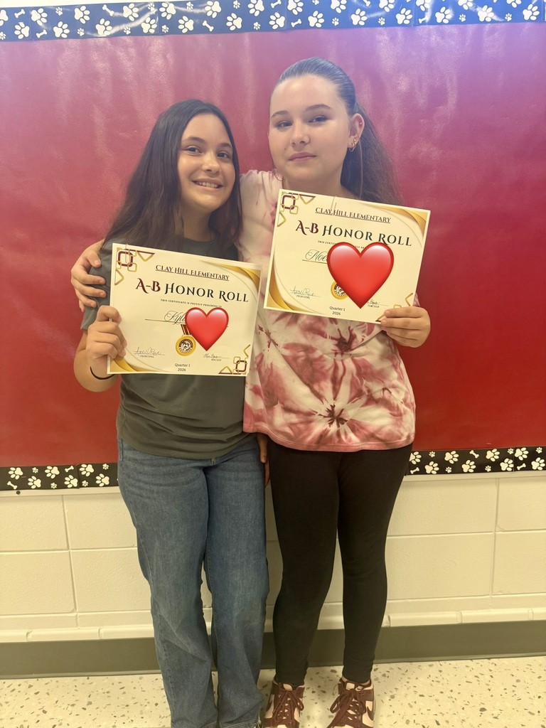Two girls hold certificates with red hearts. They stand in front of a red wall. One wears a gray top and blue jeans, and the other wears a pink top and black pants.