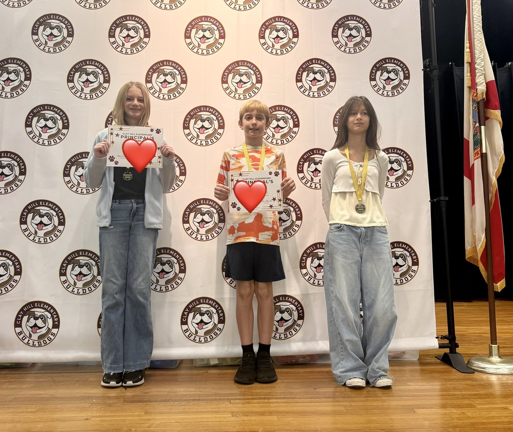 Three individuals stand on a stage, smiling. They hold heart-shaped signs. A backdrop features circular logos. A flag is partially visible.