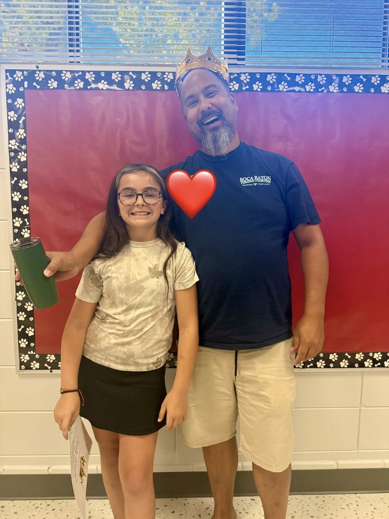 A man with a crown and a girl in glasses stand together. A red heart is in the air between them. They stand in front of a white and red wall.
