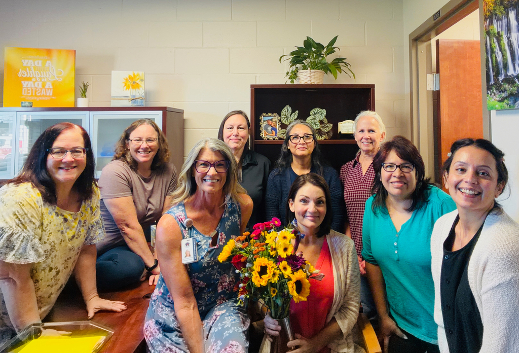 A group of women pose for a photo. One holds a bouquet of flowers. Background includes shelves with decorative items.