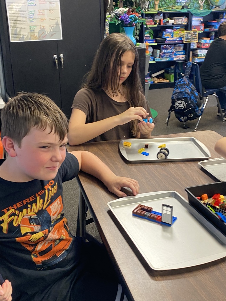 Two children work on Legos at a table. One girl sits while another boy sits. Background shows shelves and a person.