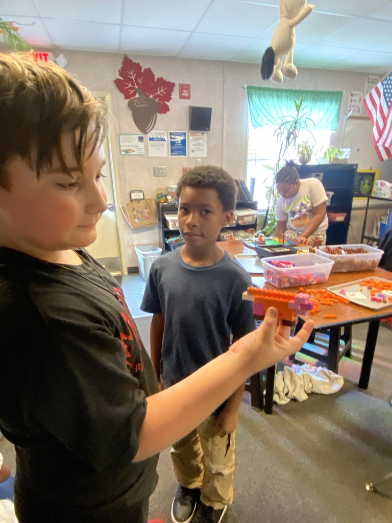 Two young boys in a classroom. One holds an object and looks at it. Behind them, a table with food, a person in a white shirt, and a flag.